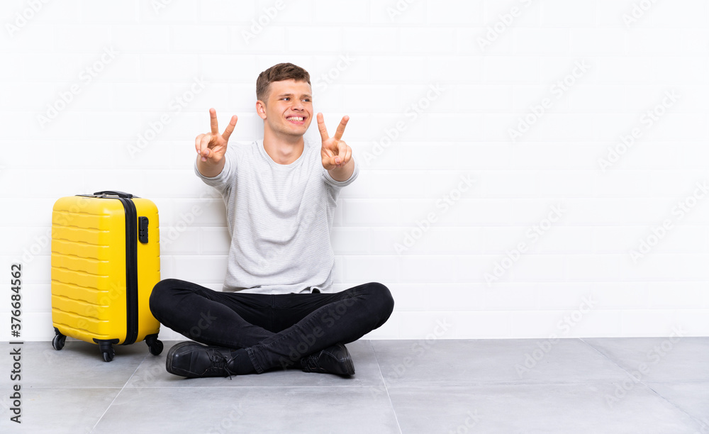 Young handsome man sitting on the floor with a suitcase smiling and showing victory sign Young handsome man sitting on the floor with a suitcase smiling and showing victory sign