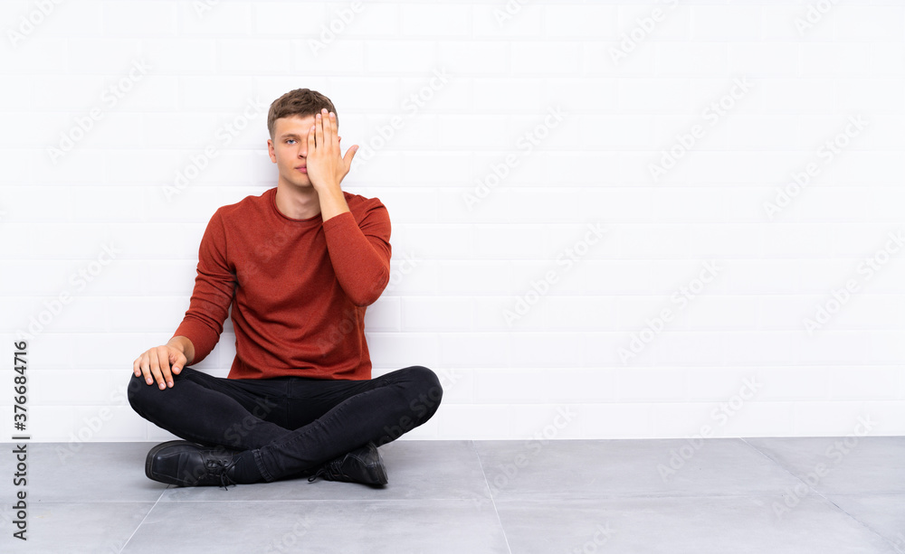 Young handsome man sitting on the floor covering a eye by hand Young handsome man sitting on the floor covering a eye by hand