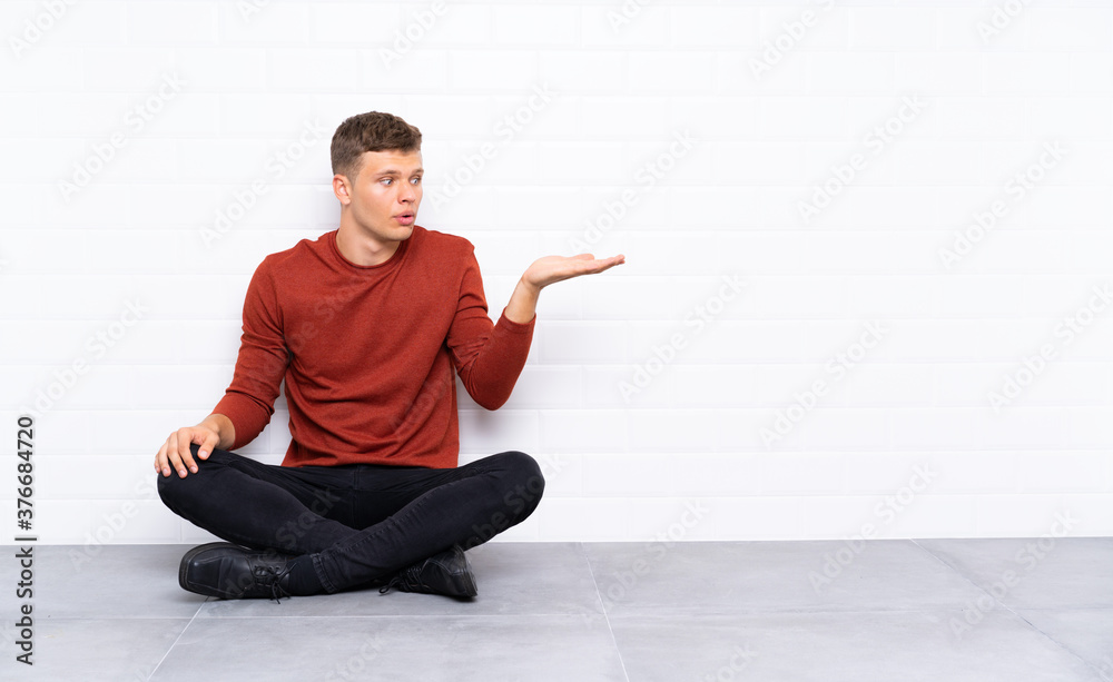 Young handsome man sitting on the floor holding copyspace imaginary on the palm Young handsome man sitting on the floor holding copyspace imaginary on the palm