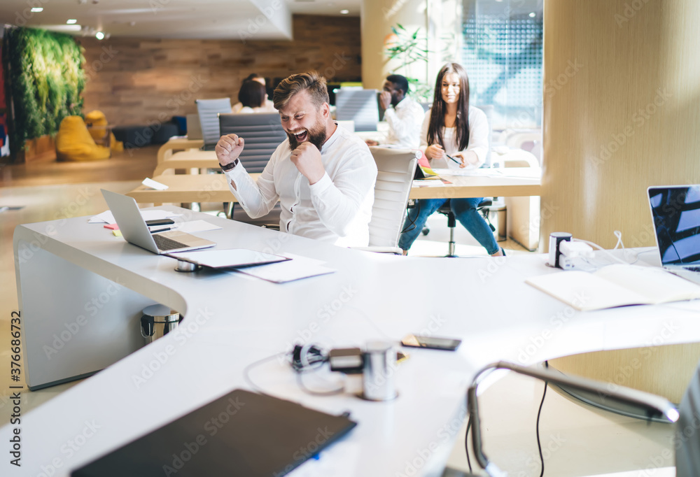 Obraz premium Emotional male sales manager celebrating accomplished startup gesture and screaming near laptop computer, expressive professional man employee excited with getting good news from exchange in office