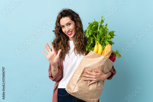 Young woman holding a grocery shopping bag isolated on blue background counting five with fingers