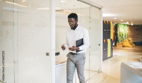 Trendy dressed african american american male employee walking in office reading information concentrated on idea, pensive dark skinned finance manager in smart casual clothes strolling on job © BullRun