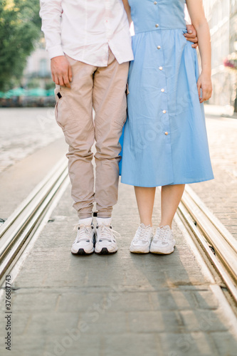 Cropped image of legs of lovely stylish young couple in love, woman in blue dress and man in beige pants, standing on the pavement road and tram track, having fun outdoors in the city