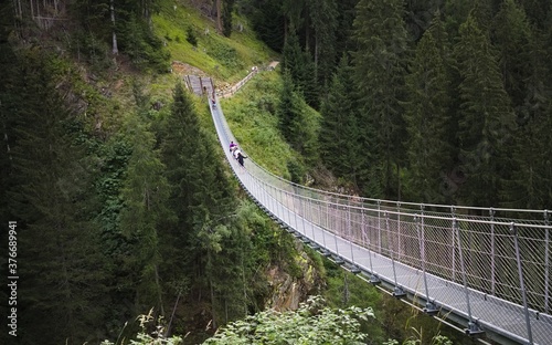 People walking on a steel tibetan bridge inside a forest in the italian Alps (Rabbi, Trentino, italy, Europe)