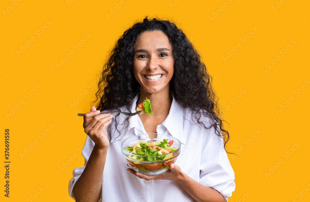 Healthy Eating. Smiling curly woman enjoying fresh vegetable salad over yellow background