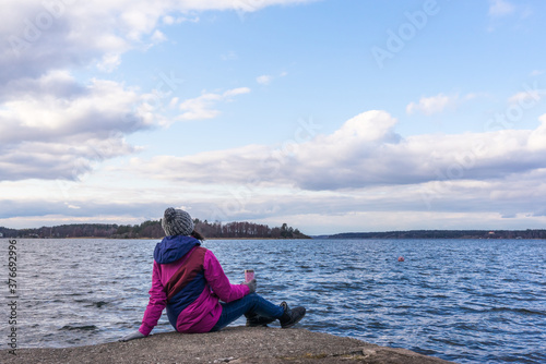 Young woman on rocky coast looking at the sea. Autumn Fall Concept. View of Scandinavian Stockholm Archipelago on cloudy sky background. Pine forest islands on the horizon.