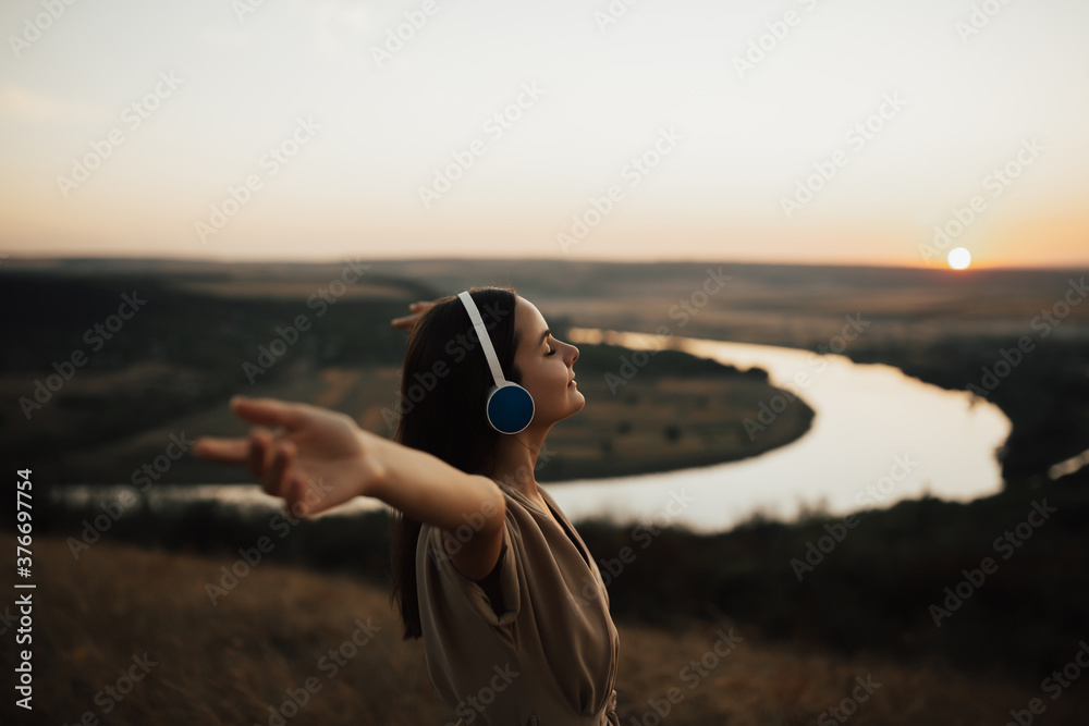 Young beautiful woman listens to music in summer during sunset. Smiling pretty woman with closed eyes and hands up feeling positive emotions. Copy space.