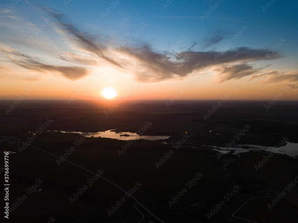 Aerial view of the sunrise over dark rural landscape with lake and river. Rising sun at blue cloudy sky over horizon and dark surface with reflecting water.