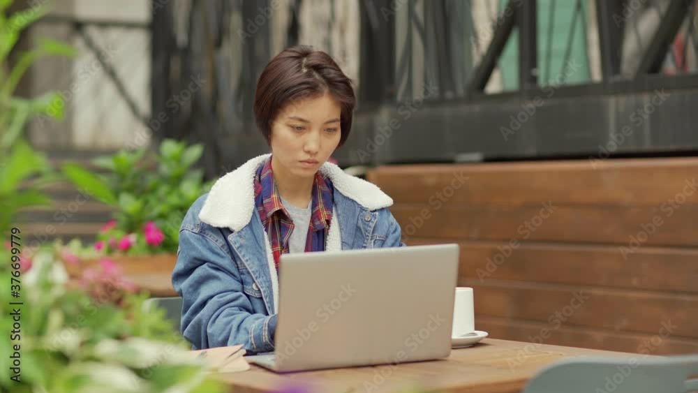 Panning zoom in shot of young Asian woman with septum nose piercing working on laptop computer, closing pc, drinking tea or coffee and thinking sitting at table at outdoor cafe
