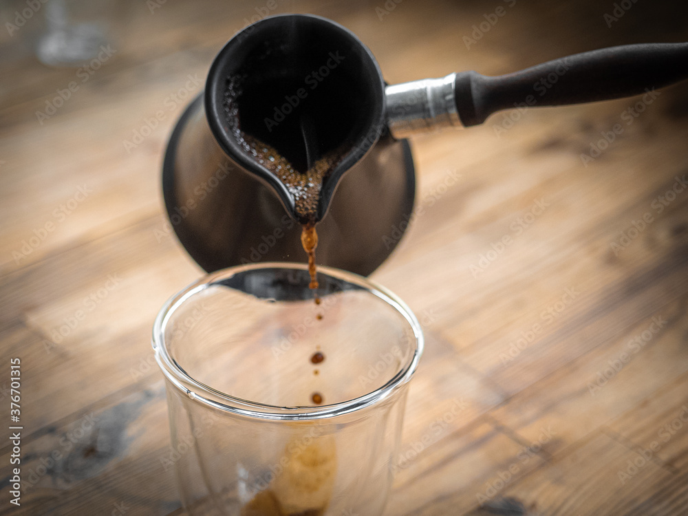 Freshly brewed turkish coffee pouring from cezve into transparent glass on wooden background. Filling hot coffee to transparent vacuum glass with double walls.