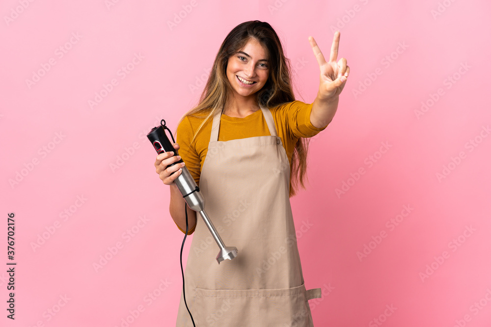 Young chef woman using hand blender isolated on pink background smiling and showing victory sign Young chef woman using hand blender isolated on pink background smiling and showing victory sign