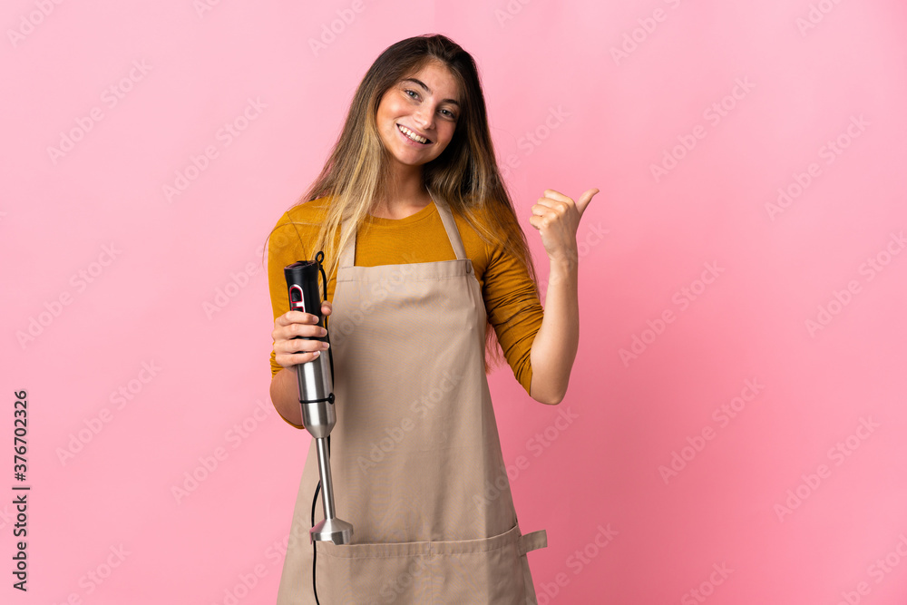 Young chef woman using hand blender isolated on pink background pointing to the side to present a product Young chef woman using hand blender isolated on pink background pointing to the side to present a product
