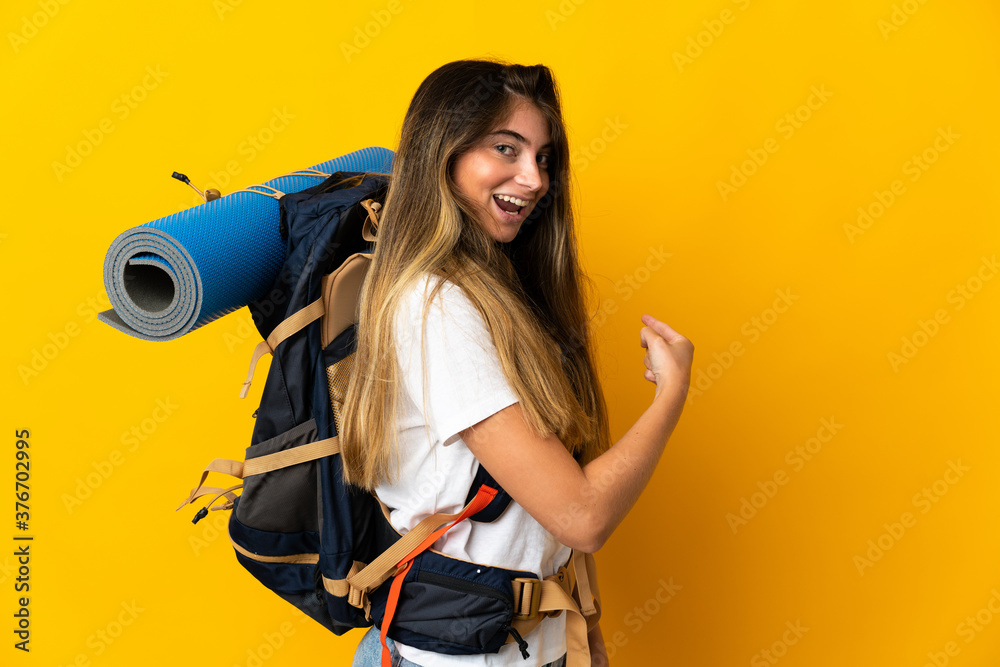 Young mountaineer woman with a big backpack isolated on yellow background pointing back Young mountaineer woman with a big backpack isolated on yellow background pointing back