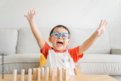 Happy smart and nerd toddler asian boy raised hand when he win play wooden toy block at home.Boy child kid wearing glasses.myopia or Short sighted.Positive human emotion, Attitude and self esteem.