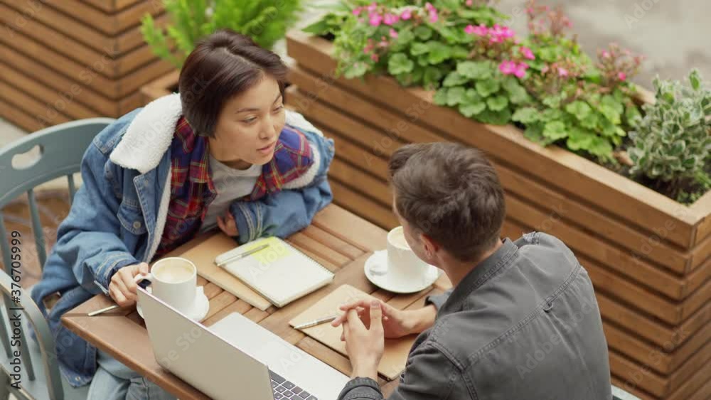 Tracking top view shot of young Asian woman and Caucasian man discussing college project or startup business ideas using laptop over coffee sitting at outdoor cafe table Tracking top view shot of young Asian woman and Caucasian man discussing college project or startup business ideas using laptop over coffee sitting at outdoor cafe table