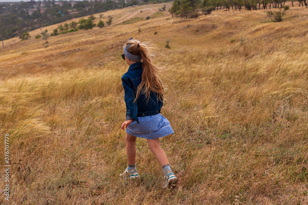 Adorable little girl in denim jacket, blue plaid dress running in yellow wild grass field. Happy stylish long blonde hair child on countryside landscape. Cute kid walking outdoor rural road trip.