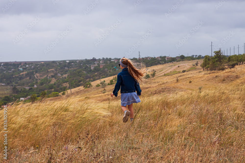 Adorable little girl in denim jacket, blue plaid dress running in yellow wild grass field. Happy stylish long blonde hair child on countryside landscape. Cute kid walking outdoor rural road trip.