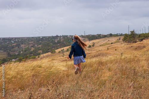 Adorable little girl in denim jacket  blue plaid dress running in yellow wild grass field. Happy stylish long blonde hair child on countryside landscape. Cute kid walking outdoor rural road trip.