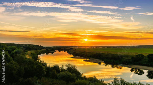 Scenic view at beautiful summer river sunset with reflection on water with green bushes  grass  golden sun rays  calm water  deep blue cloudy sky and glow on a background  spring evening landscape