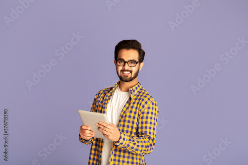Online work, education or communication. Funky Indian man using tablet computer on violet background