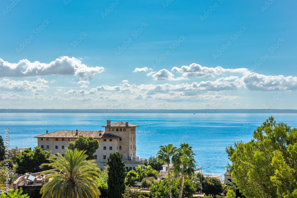 Building in front of the sea with trees in the foreground, in the background views of the sea and the blue sky with white clouds