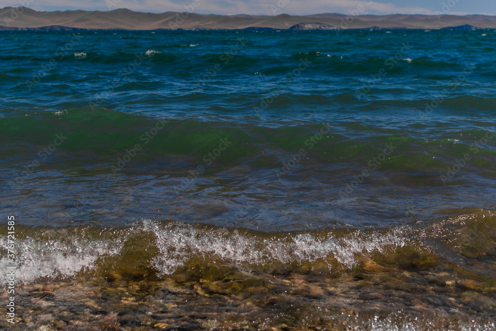 bright blue emerald green waves of lake baikal in the light of the sun in summer, mountains on the horizon, stones in water, coast bright blue emerald green waves of lake baikal in the light of the sun in summer, mountains on the horizon, stones in water, coast