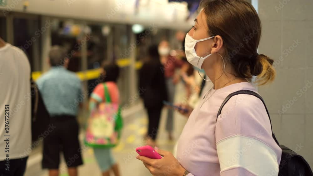 Young woman wearing white medical face mask waiting train at the station. Casual woman using smart phone in train station Young woman wearing white medical face mask waiting train at the station. Casual woman using smart phone in train station