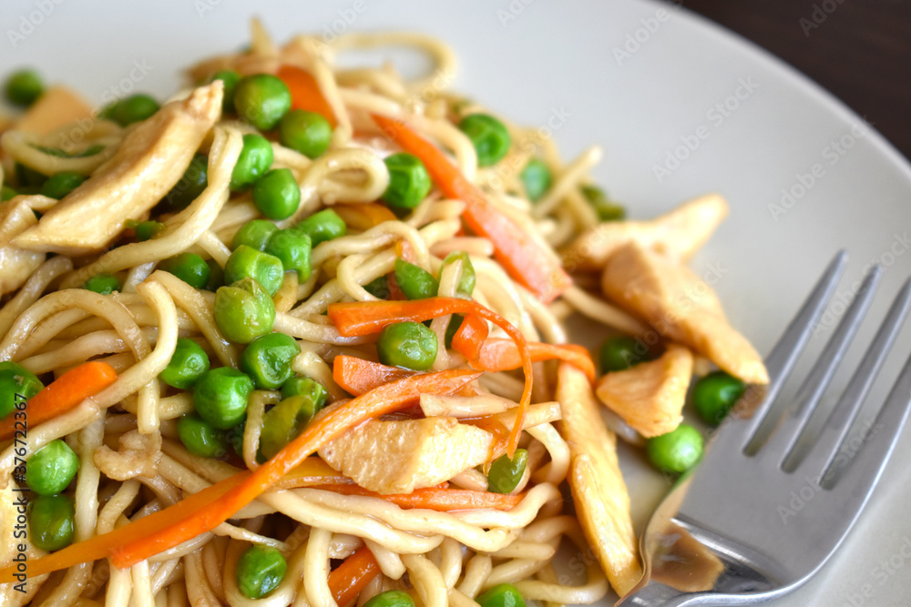 Chicken chow mein on a white plate with a fork on the right side. Stir fry noodles with chicken, carrot and green peas. Chicken chow mein on a white plate with a fork on the right side. Stir fry noodles with chicken, carrot and green peas.
