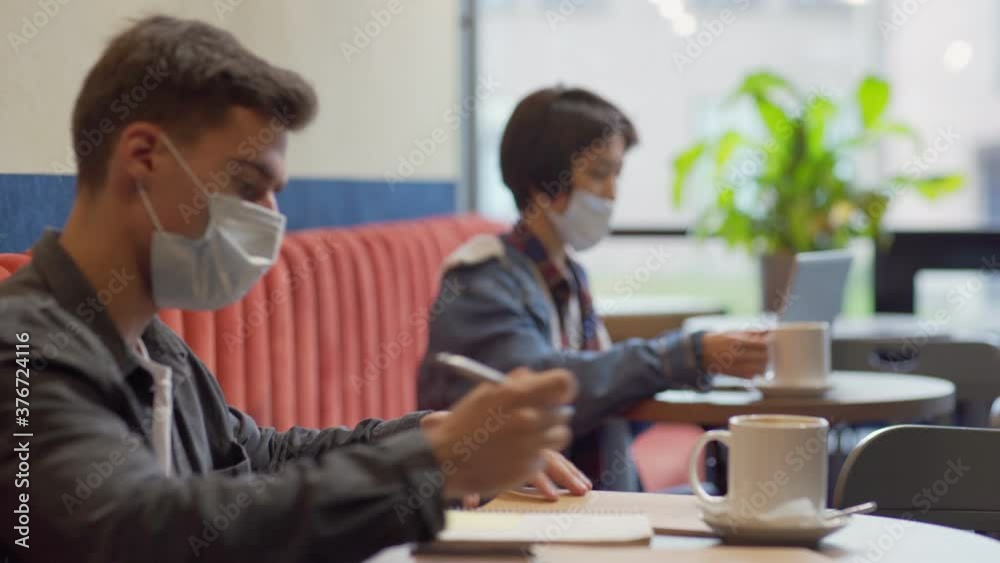 Camera moving from young Asian woman working on laptop computer and having coffee to man making notes in notepad sitting at table in cafe, both wearing face masks Camera moving from young Asian woman working on laptop computer and having coffee to man making notes in notepad sitting at table in cafe, both wearing face masks