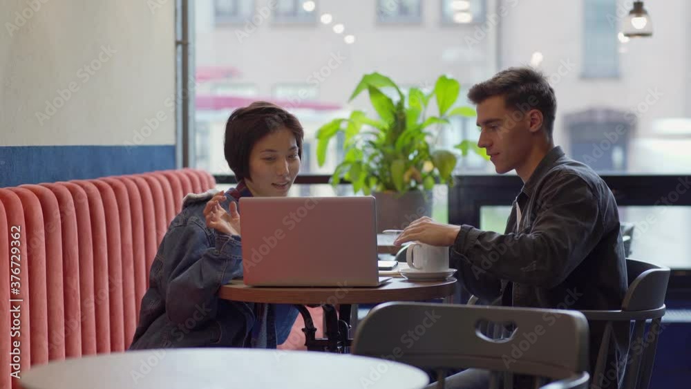 Tilt down side view of diverse young man and woman looking at laptop screen and discussing startup business ideas over coffee at cafe table Tilt down side view of diverse young man and woman looking at laptop screen and discussing startup business ideas over coffee at cafe table