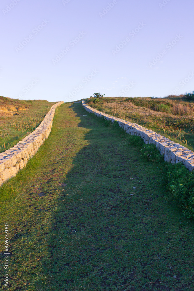 Camino de hierba bordeado por muretes de piedra que se aleja hacia un cielo despejado. Iluminación lateral.