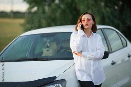 beautiful young woman in pink glasses with mobile phone near car