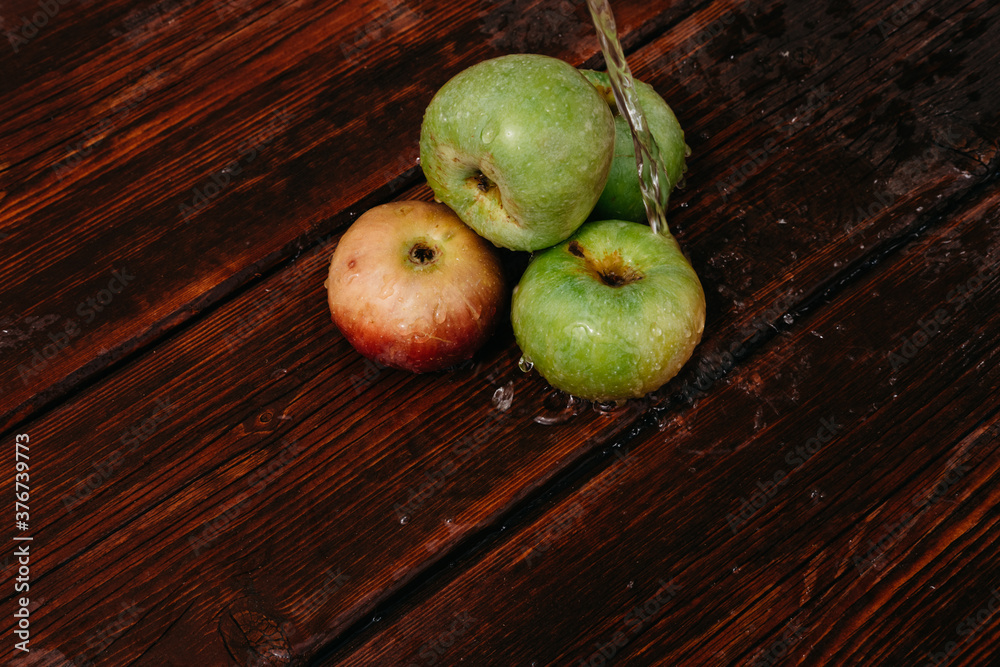 Splashes of clean water pour over freshly picked apples of the autumn harvest. Red and green apples lie on the mahogany wooden kitchen table, and water drips from the top. Splashes of clean water pour over freshly picked apples of the autumn harvest. Red and green apples lie on the mahogany wooden kitchen table, and water drips from the top.