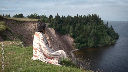 Lonely woman in felt hat and woolen poncho sits on grassy seashore and looks at wooded cape. Travel, adventure, feeling nature, folk ethnic lifestyle. Lake Onega, Russian North.
