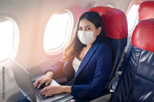 A young businesswoman wearing face mask is using laptop onboard, New normal travel after covid-19 pandemic concept