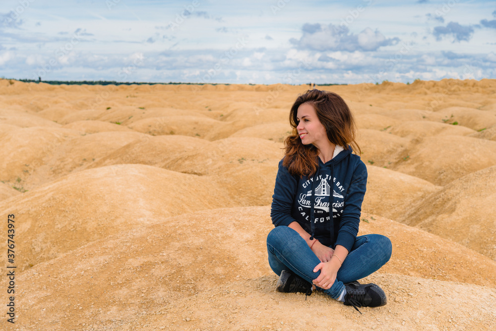 Beautiful young woman in a sweatshirt with an inscription sits on the sand in the desert with sand dunes of bizarre shape