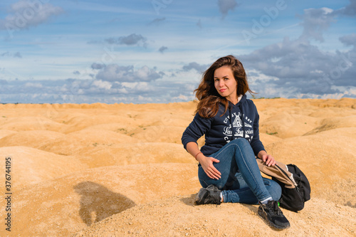 Beautiful young woman in a sweatshirt with an inscription sits on the sand in the desert with sand dunes of bizarre shape © KseniaJoyg