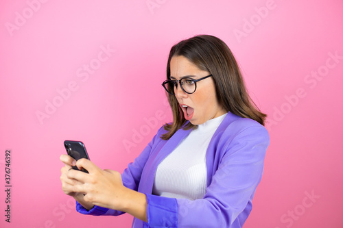 Young beautiful business woman over isolated pink background chatting with her phone and surprised