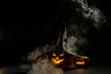 Three jack o lanterns glow in the dark amidst the fog. Halloween pumpkin in a witch hat.