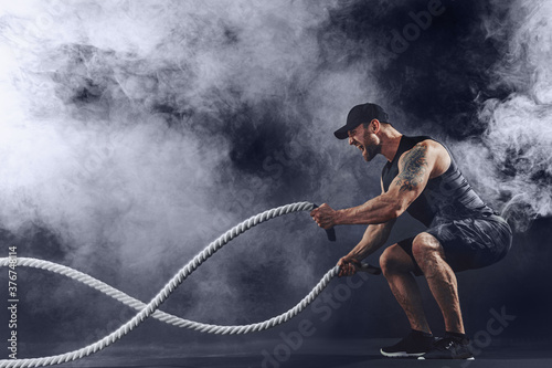 Tablou canvas Bearded athletic looking bodybulder work out with battle rope on dark studio background with smoke