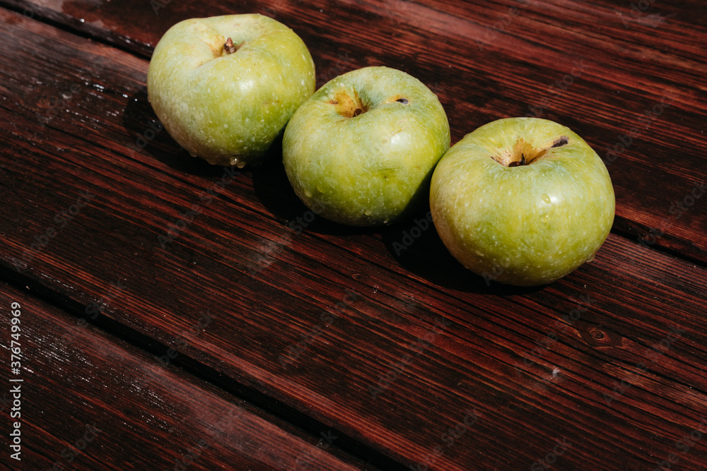 Fresh autumn Apple harvest on the kitchen countertop. Three fresh green apples lie in a row on a wooden table. A textured mahogany wood background and fruit lie on it. Fresh autumn Apple harvest on the kitchen countertop. Three fresh green apples lie in a row on a wooden table. A textured mahogany wood background and fruit lie on it.