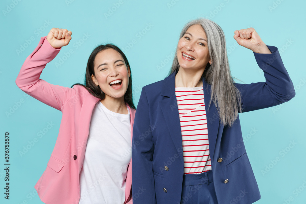 Happy two asian female business women employer and employee in pink blue jackets posing working in office clenching fists doing winner gesture isolated on pastel blue color background studio portrait. Happy two asian female business women employer and employee in pink blue jackets posing working in office clenching fists doing winner gesture isolated on pastel blue color background studio portrait.