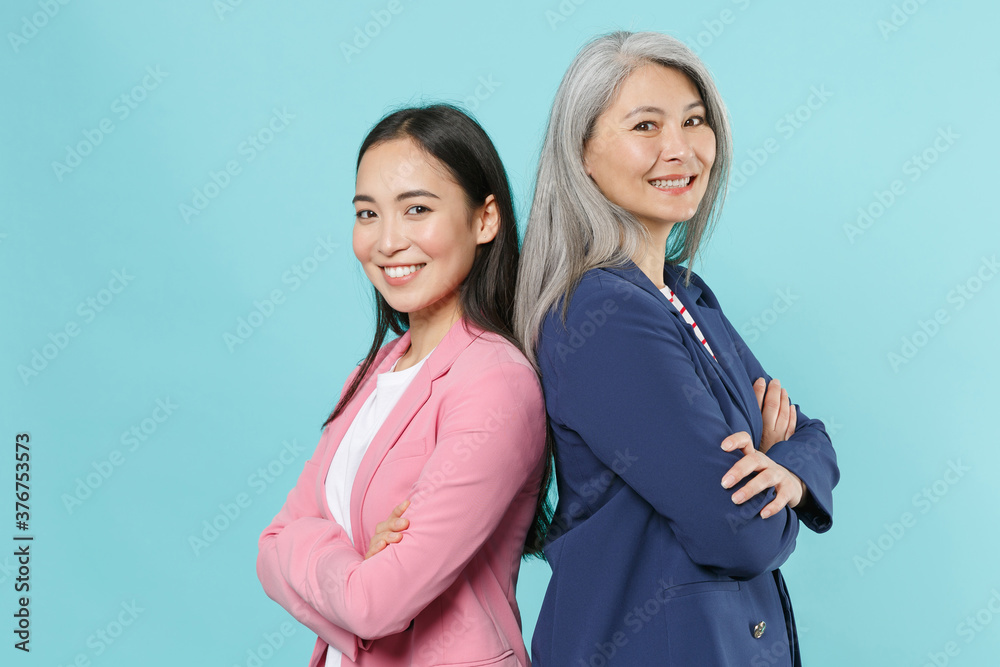 Smiling two asian female business women employer employee in pink blue jackets working in office stand back to back hold hands crossed looking camera isolated on blue color background studio portrait. Smiling two asian female business women employer employee in pink blue jackets working in office stand back to back hold hands crossed looking camera isolated on blue color background studio portrait.