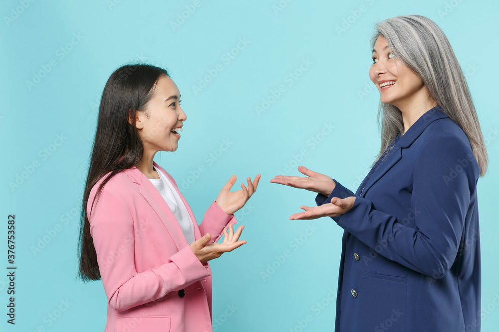 Side view of cheerful funny two asian female business women employer employee in pink blue jackets working in office speaking talking spreading hands isolated on blue color background studio portrait. Side view of cheerful funny two asian female business women employer employee in pink blue jackets working in office speaking talking spreading hands isolated on blue color background studio portrait.