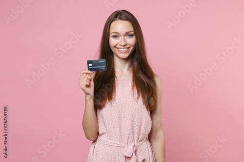 Smiling beautiful attractive young brunette woman 20s wearing pink summer dotted dress posing holding in hand credit bank card looking camera isolated on pastel pink color background studio portrait.