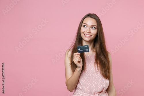 Smiling beautiful pensive young brunette woman 20s wearing pink summer dotted dress posing holding in hand credit bank card looking up isolated on pastel pink color background studio portrait.