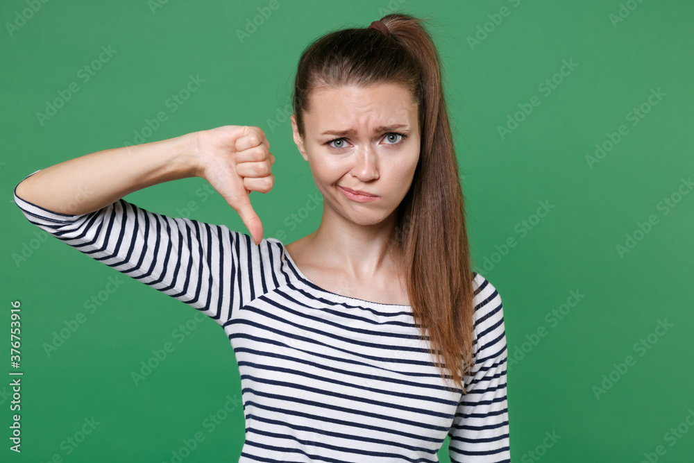 Displeased dissatisfied confused puzzled young brunette woman 20s wearing striped casual clothes posing standing showing thumb down looking camera isolated on green color background, studio portrait. Displeased dissatisfied confused puzzled young brunette woman 20s wearing striped casual clothes posing standing showing thumb down looking camera isolated on green color background, studio portrait.