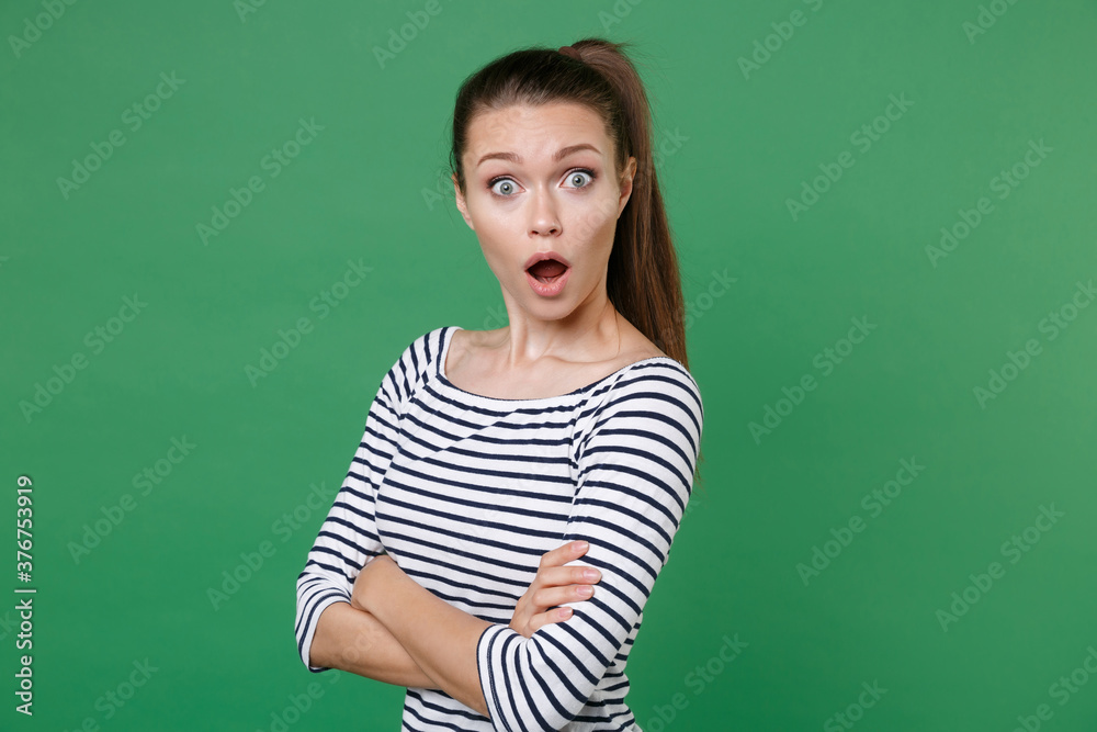 Shocked worried nervous young brunette woman 20s wearing striped casual clothes posing stand holding hands crossed keeping mouth open looking camera isolated on green color background studio portrait. Shocked worried nervous young brunette woman 20s wearing striped casual clothes posing stand holding hands crossed keeping mouth open looking camera isolated on green color background studio portrait.