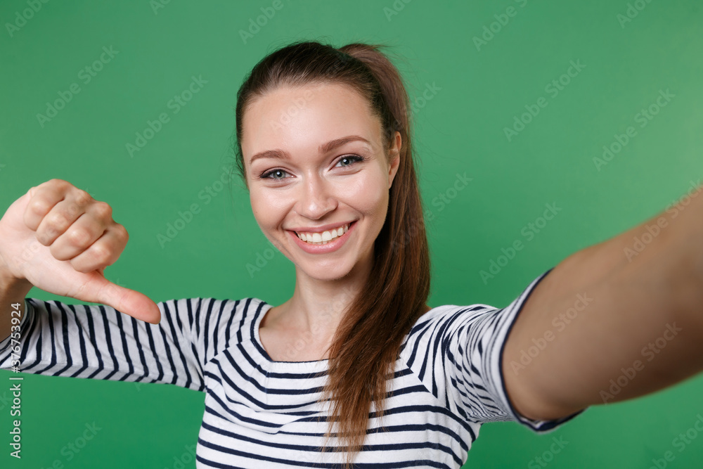 Close up of smiling young brunette woman 20s wearing striped casual clothes posing doing selfie shot on mobile phone pointing thumb on herself isolated on green color wall background studio portrait. Close up of smiling young brunette woman 20s wearing striped casual clothes posing doing selfie shot on mobile phone pointing thumb on herself isolated on green color wall background studio portrait.