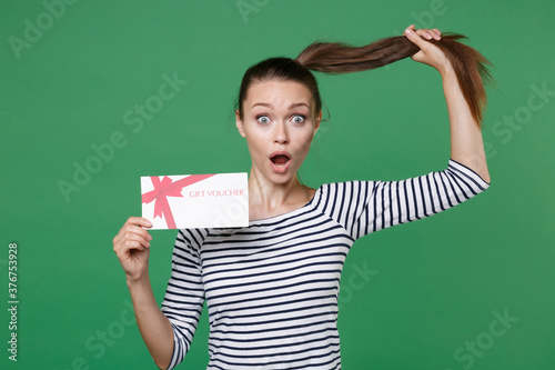 Shocked amazed young brunette woman 20s in striped casual clothes posing holding in hand gift certificate hold ponytail keeping mouth open looking camera isolated on green background studio portrait.